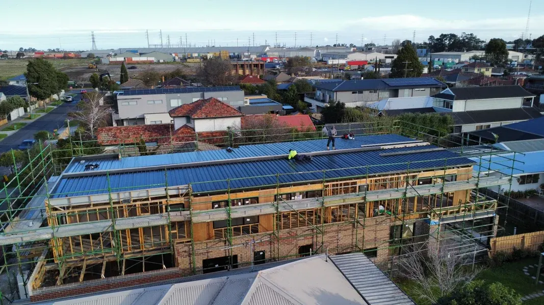 Drone view of a scaffolded double-storey townhouse block getting a new blue metal roof, roofers up top, with tiled houses around it and an industrial skyline behind