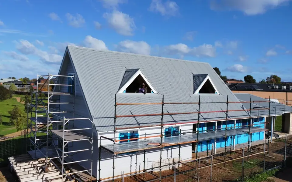 A large gable house in scaffolding under a blue sky, its new light grey metal roof finished around two triangular dormer windows