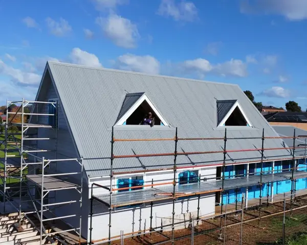 A large gable house in scaffolding under a blue sky, its new light grey metal roof finished around two triangular dormer windows
