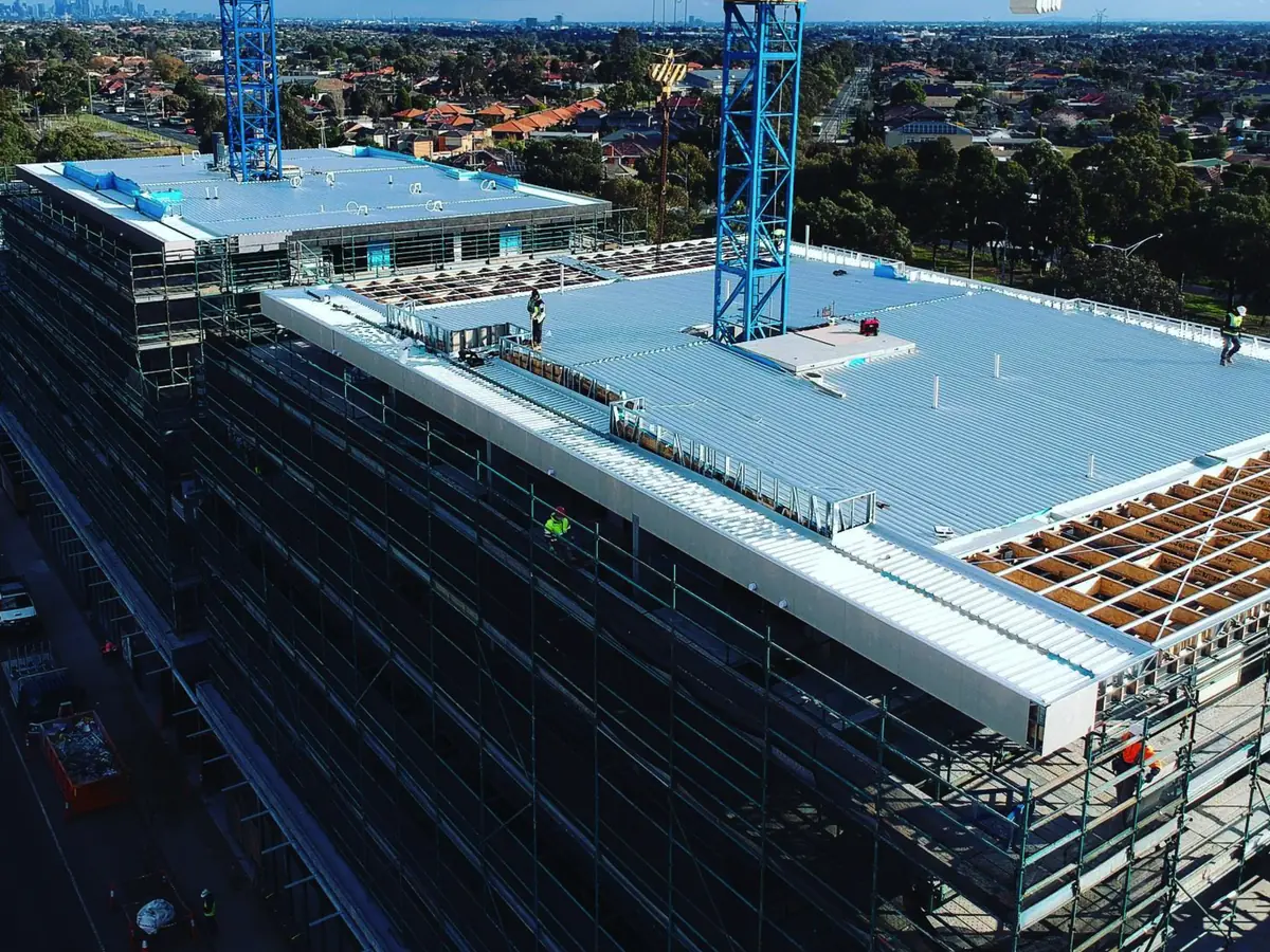 Drone view of a multi-storey building in scaffolding with a large low-pitch metal roof being laid, roofers in hi-vis on top, a tower crane, and suburbs and the city skyline behind