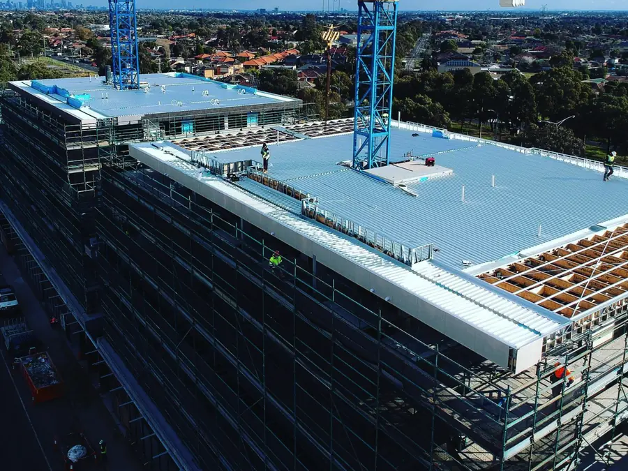 Drone view of a multi-storey building in scaffolding with a large low-pitch metal roof being laid, roofers in hi-vis on top, a tower crane, and suburbs and the city skyline behind