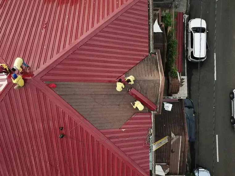 Top-down drone view of two roofers working on a red metal roof beside a suburban street