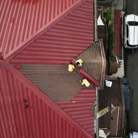 Top-down drone view of two roofers working on a red metal roof beside a suburban street