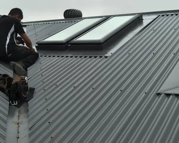 A roofer kneeling at the ridge of a dark grey corrugated metal roof, beside two skylights and a whirlybird vent