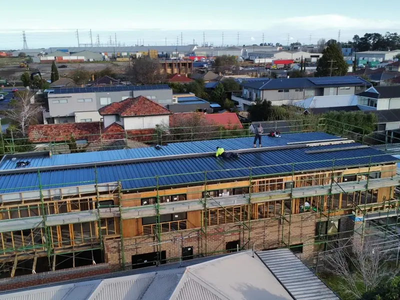 Drone view of a long double-storey townhouse block in scaffolding, its new dark metal roof going on with two roofers up top