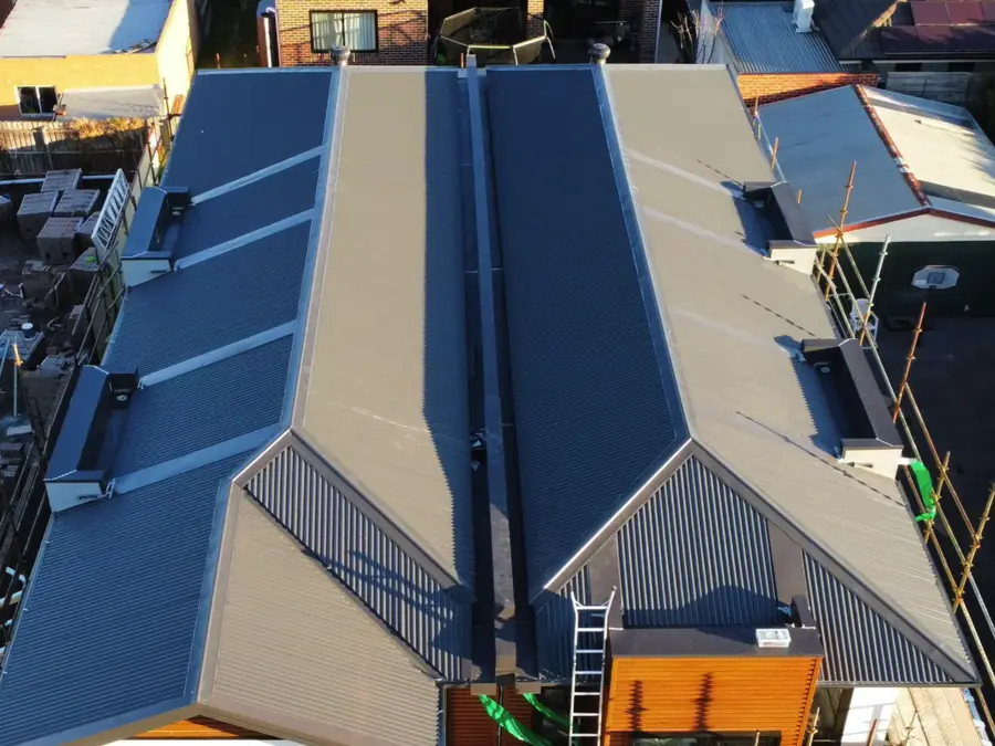 Drone view looking down on a pair of twin-gable townhouses with new dark grey corrugated metal roofs, a ladder and scaffolding at the front
